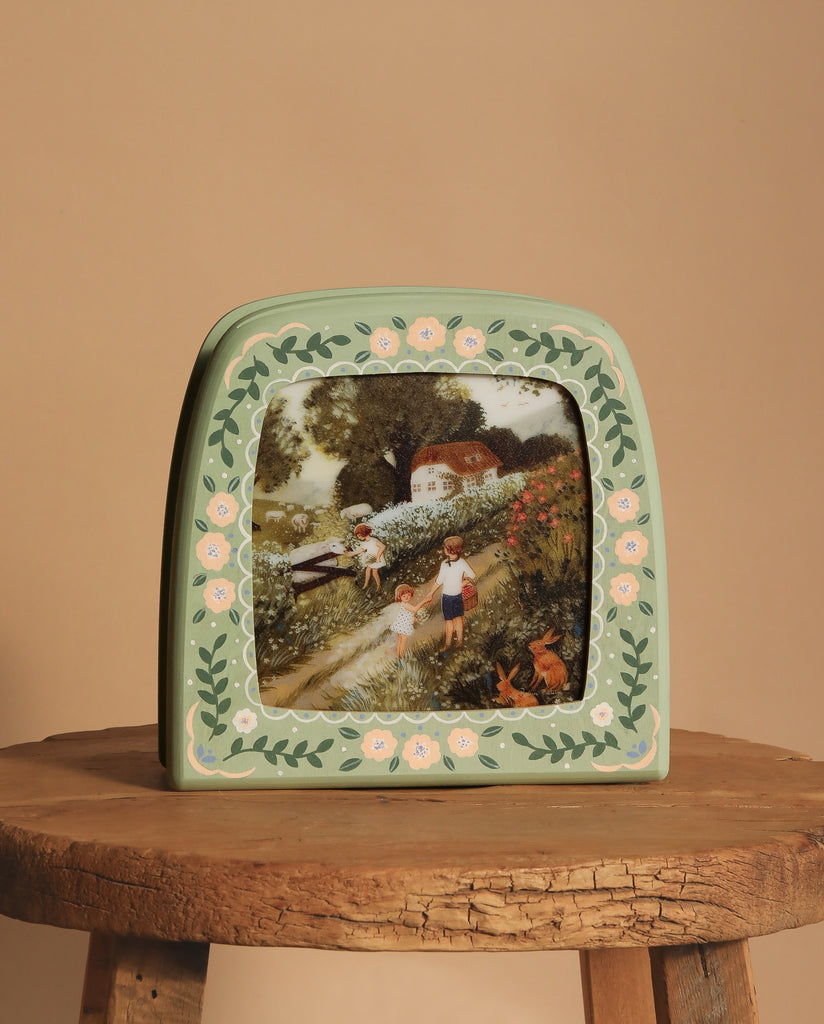 A framed picture sits on a round wooden table beside the Hand-Painted Toverlux Magic Lamp - Green Meadow. The frame, with floral designs, encloses an image of two children and a dog on a countryside path. The lamp's soft glow enchants the scene against the neutral beige background.