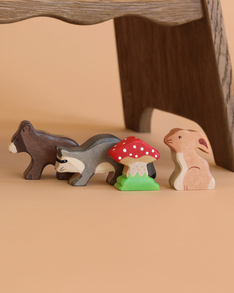 The Holztiger Handmade Wooden Woodland Animals Set features a bear, badger, rabbit, and a red-and-white mushroom displayed in a row on a tan background beneath a wooden surface.