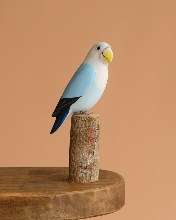 The Hand Carved Ornament - Lovebird - Light Blue - 7.5"H features a hand-painted, eco-friendly figurine perched on sustainable softwood, displayed on a wooden table with a plain peach background.