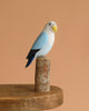 The Hand Carved Ornament - Lovebird - Light Blue - 7.5"H features a hand-painted, eco-friendly figurine perched on sustainable softwood, displayed on a wooden table with a plain peach background.