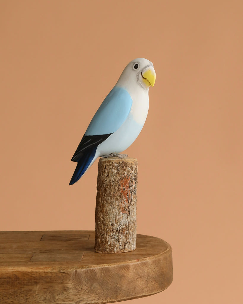 The Hand Carved Ornament - Lovebird - Light Blue - 7.5"H features a hand-painted, eco-friendly figurine perched on sustainable softwood, displayed on a wooden table with a plain peach background.