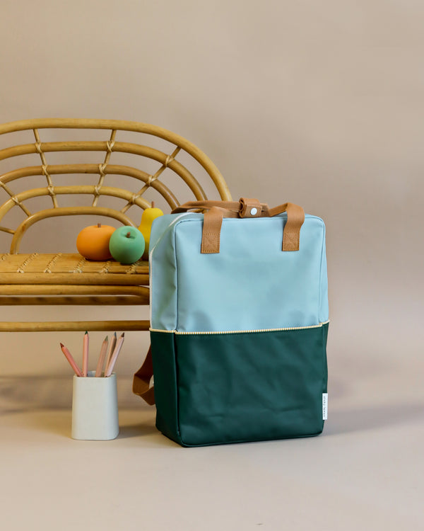 A stylish Sticky Lemon Backpack Large in island blue and dark teal, crafted from waterproof nylon, sits on the floor against a beige background, beside a wicker chair with fruit and a mug on it.