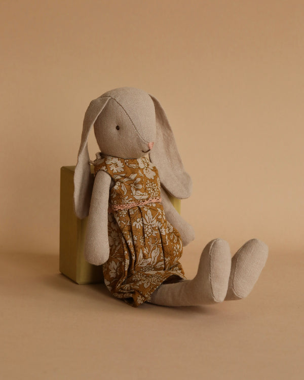 A soft beige Maileg Bunny, Size 2 - Flower Dress with long floppy ears is sitting against a light-brown background. Made from soft natural fabrics and recycled polyester, the bunny is wearing a brown floral dress and leaning slightly to one side, with its legs stretched out in front.
