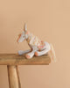 A soft, pastel-colored unicorn plush toy with a yarn mane and tail sits on a rustic wooden stool against a plain beige background, evoking the whimsical charm of the Maileg Fairytale Set.