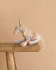 The Maileg Unicorn, Small—a soft toy with a gray body, cream mane and tail, golden horn, and decorative saddle—sits on a rustic wooden stool against a beige background.