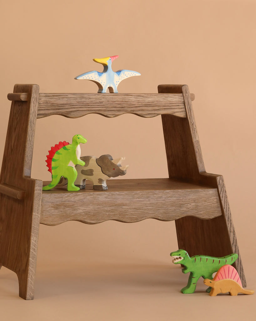 A small wooden shelf displays the Holztiger Handmade Wooden Dinosaur Set, featuring a pterodactyl on top, green and red stegosaurus, gray triceratops, green T-rex, and orange stegosaurus at the bottom.