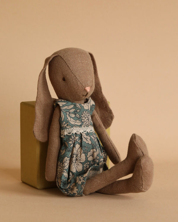 A brown Maileg Bunny, Size 1, Brown - Blossom Dress leans against a yellow-green box. The beige background complements the soft, neutral, and cozy aesthetic of the FW23 collection scene.