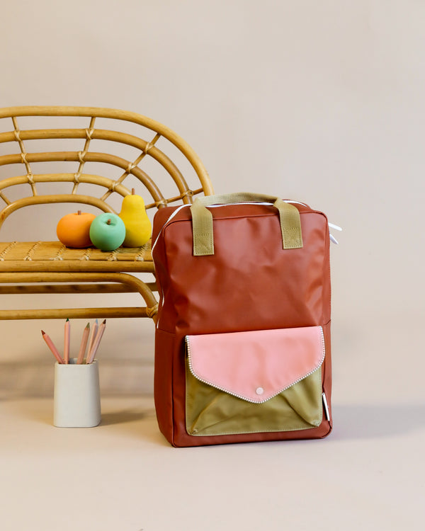 A Lighthouse Red Sticky Lemon backpack made from recycled PET bottles with a pink flap and olive green details stands upright against a rattan chair. On the chair lies a notepad and two colorful fruit-shaped stress balls, next to it.