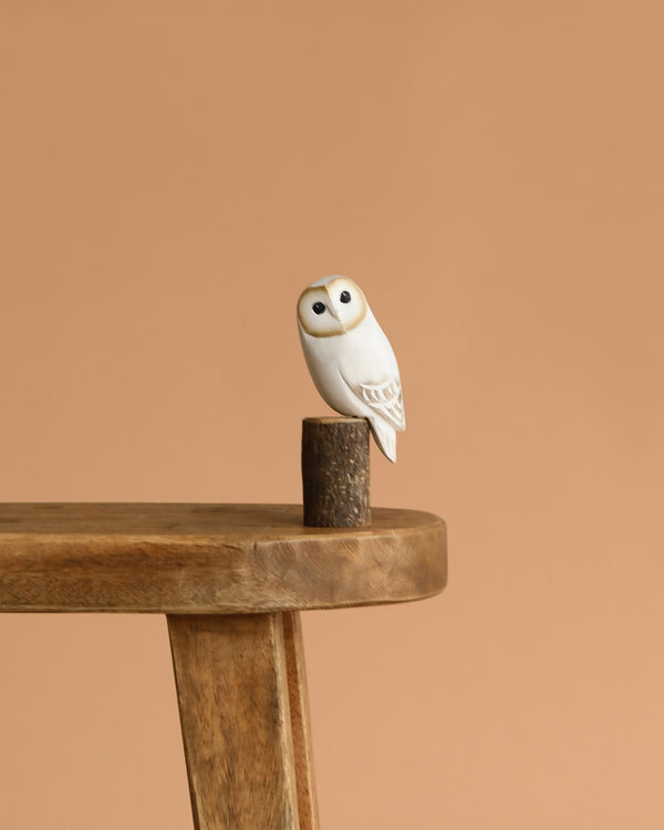 The Hand Carved Ornament - Mini Barn Owl - 5"H rests on a round wooden pedestal atop a stool against a plain beige background—an ideal sustainable decor piece for any space.