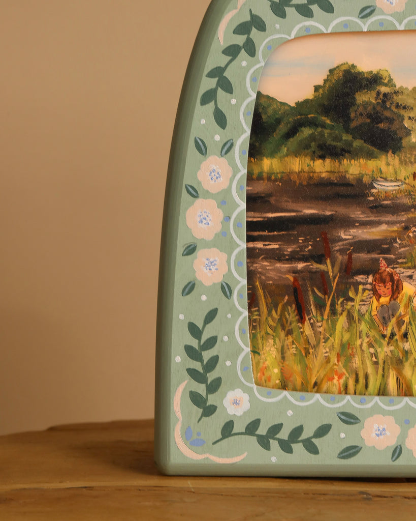 A decorative box, painted with a nature scene of lush greenery and blooming flowers, mirrors the elegance of the Hand-Painted Toverlux Magic Lamp - Green Meadow. It's placed on a wooden surface with intricate floral patterns framing the scene.