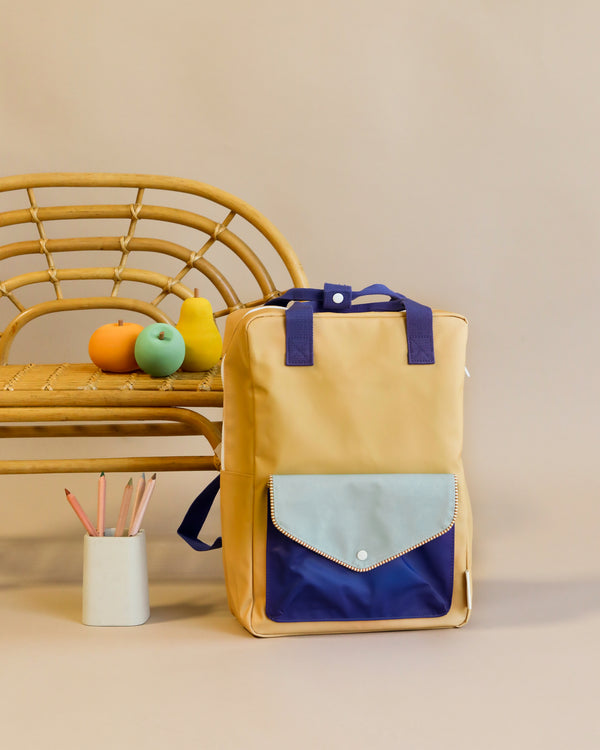 A stylish Camp Yellow Sticky Lemon Backpack Large with a flap closure sits in front of a bamboo chair. Next to the chair, a white cup holds pencils and a trio of colorful toy fruits rests on the seat.