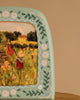 A green frame with floral patterns surrounds a painting of children playing in a field, one raising a red flag, another sitting near trees and a yellow house under the clear sky—reminiscent of scenes on the Hand-Painted Toverlux Magic Lamp - Green Meadow.