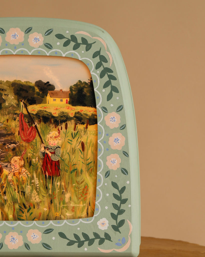 A green frame with floral patterns surrounds a painting of children playing in a field, one raising a red flag, another sitting near trees and a yellow house under the clear sky—reminiscent of scenes on the Hand-Painted Toverlux Magic Lamp - Green Meadow.