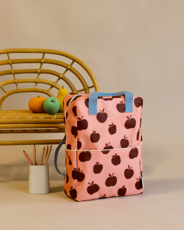 A peach-colored Sticky Lemon Backpack Large | Special Edition with a black apple pattern, made from recycled PET bottles, stands against a gray backdrop beside a bamboo chair and a jar with art supplies.
