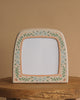 A square mirror with a rounded frame, featuring a hand-painted floral pattern in green and orange hues, echoes the charm of Magic Silhouettes. Its delicate leaves and flowers have a scalloped edge and is set on a rustic wooden table against a warm beige background.