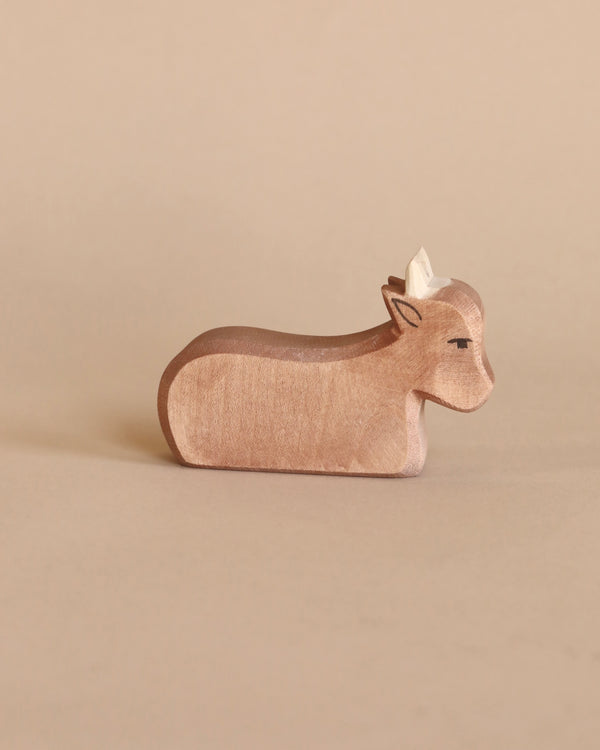 A handcrafted Ostheimer Ox figurine with visible wood grain, set against a plain, light beige background. The ox is lying down and has a calm expression.