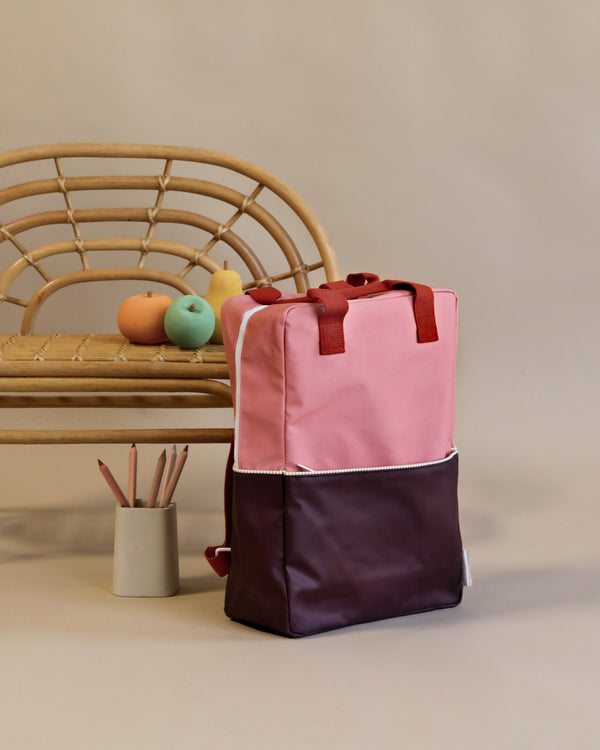 A Sticky Lemon Backpack Large | Color Blocking | Meet Me In The Meadows | Moonrise Pink with a YKK zipper stands on a floor against a beige background, next to a wooden bench with a duck and a ball on it, and a holder with coloring pencils.