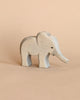 The Ostheimer Handmade Wooden Safari Animals Set features a small, light gray handcrafted wooden elephant with simple carved details, standing on a beige surface—perfect for imaginative play.