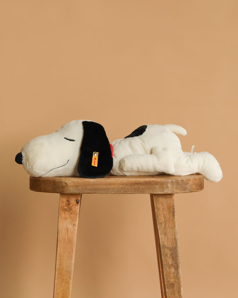 The Steiff XL Snoopy Lying Down 16" toy rests on its side atop a wooden stool set against a plain beige background.