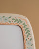 Close-up of a Hand-Painted Toverlux Magic Lamp - Spring, an Odin Parker exclusive, showcasing blue flowers and green leaves on a beige frame with light orange borders. The enchanting piece is beautifully complemented by its soft brown background.