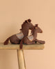 Two Maileg Pony, Small stuffed horses with patterned saddle cloths, made from cotton linen and filled with recycled polyester, sit on a rustic wooden bench against a plain beige background.