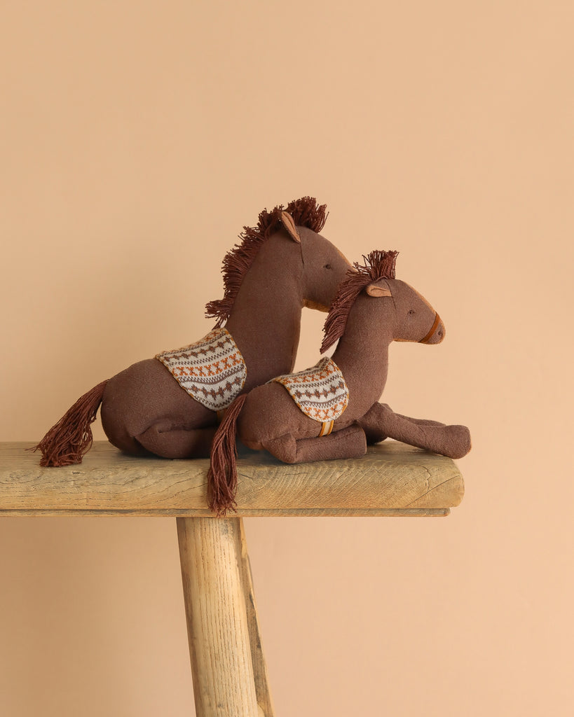 Two Maileg Pony, Small stuffed horses with patterned saddle cloths, made from cotton linen and filled with recycled polyester, sit on a rustic wooden bench against a plain beige background.