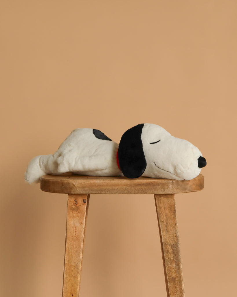 The Steiff XL Snoopy Lying Down 16", featuring black ears and a black spot on its back, is resting on its side atop a wooden stool with a plain beige background.