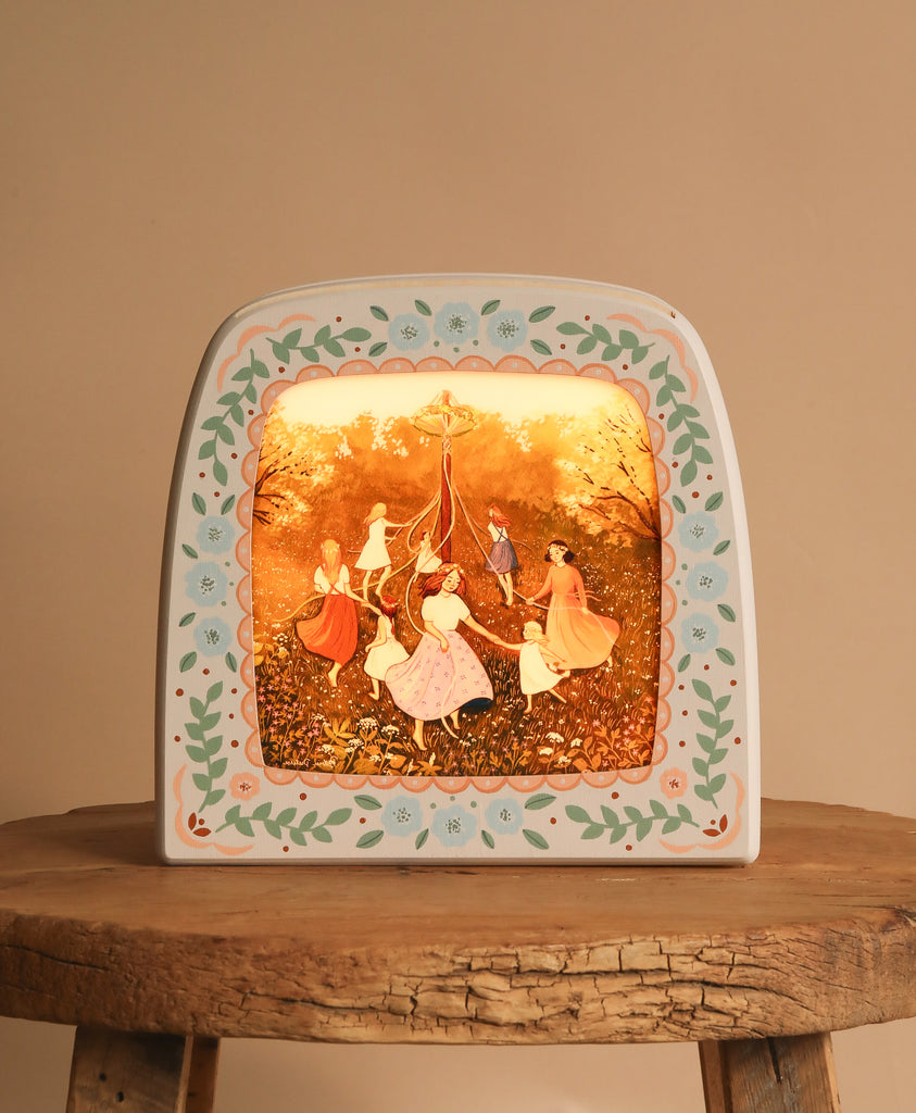 A decorative, Hand-Painted Toverlux Magic Lamp - Spring (Odin Parker Exclusive) frame, with floral borders on a wooden surface, showcases silhouettes of people in traditional attire dancing around a maypole in a lush, golden-hued setting.