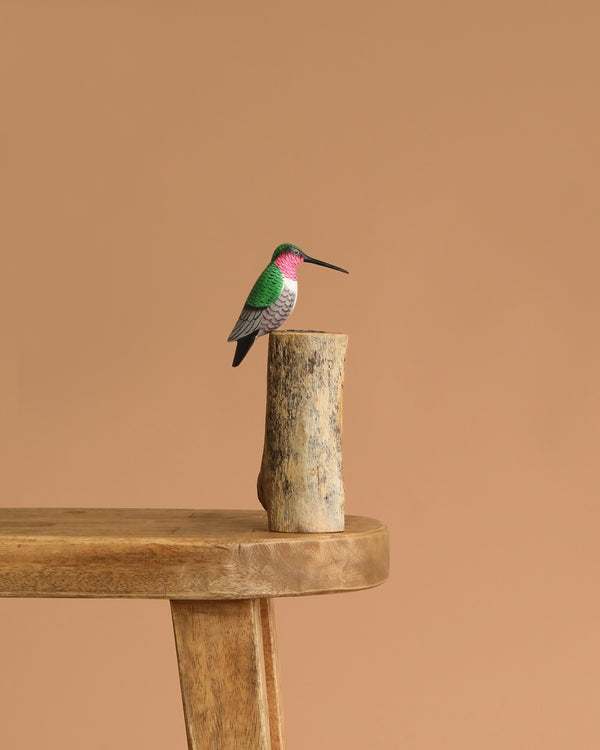 The Hand Carved Ornament - Humming Bird - 7.5"H features a small, green, white, and pink humming bird figurine perched on a natural wood base placed on a wooden table with a plain beige backdrop.