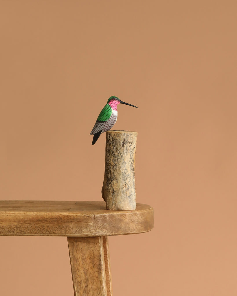 The Hand Carved Ornament - Humming Bird - 7.5"H features a small, green, white, and pink humming bird figurine perched on a natural wood base placed on a wooden table with a plain beige backdrop.