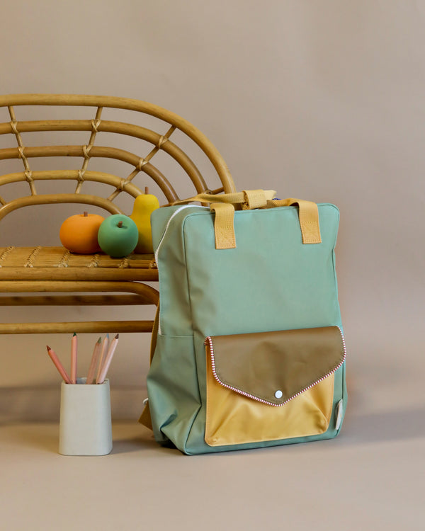 A mint green Sticky Lemon Backpack Large from the Envelope Collection in Map Green with a front pocket is resting against a rattan chair. Next to it, there is a holder with colored pencils and two fruit-shaped toys on the chair.