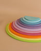 A set of colorful, semi-circular wooden stacking toys, reminiscent of modern architecture, is arranged in a gradient from largest to smallest. The pieces are in various shades of green, yellow, orange, red, pink, purple, and blue. The background is a plain beige surface.