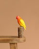 The Hand Carved Ornament - Lovebird - 7.5"H features an orange-headed, yellow-winged bird perched on a small wooden stump, displayed on a wooden bench with a plain beige background.
