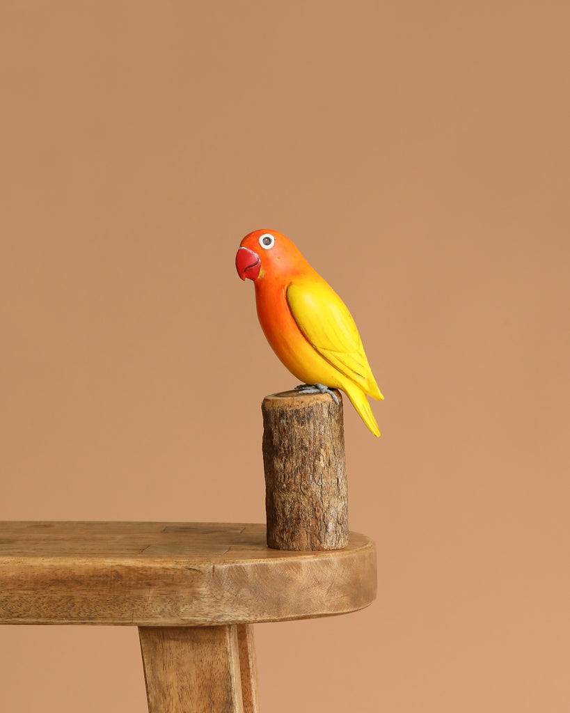 The Hand Carved Ornament - Lovebird - 7.5"H features an orange-headed, yellow-winged bird perched on a small wooden stump, displayed on a wooden bench with a plain beige background.