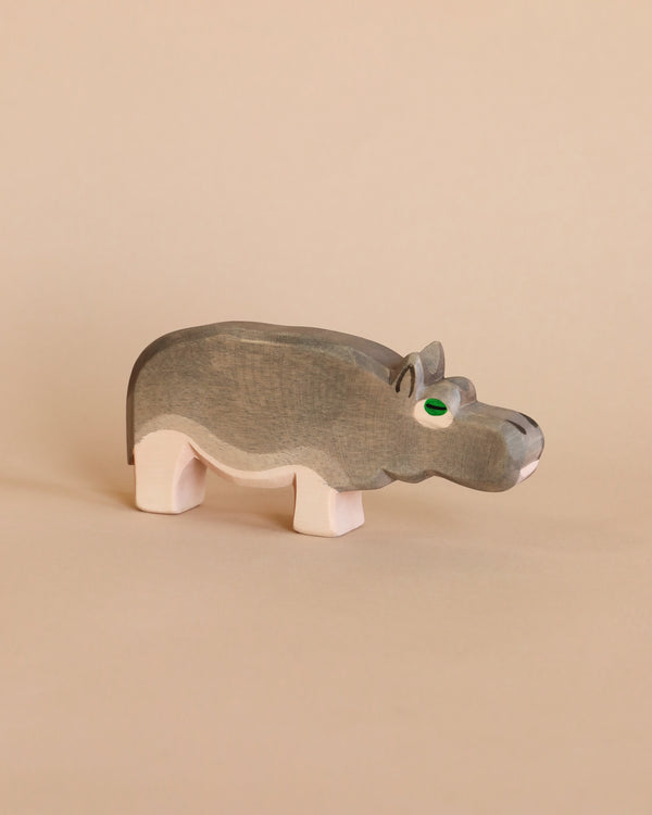 A handcrafted Ostheimer Hippopotamus figurine with green eyes stands against a plain beige background. The toy is simple, with smooth lines and visible wood grain.