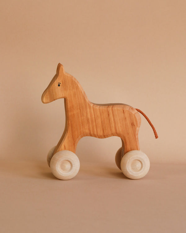 A Grimm's Wooden Horse Willy Push Toy with simple features and four white wheels stands against a neutral beige background. Made in Germany, the push toy has a smooth finish, a small visible eye, and a brown tail made of a thin strip of non-toxic material.