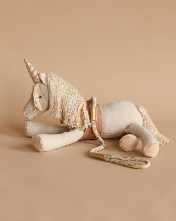 A Maileg Unicorn made of recycled polyester with a colorful mane and tail, a golden horn, and a painted wooden flute beside it, set against a soft beige background.