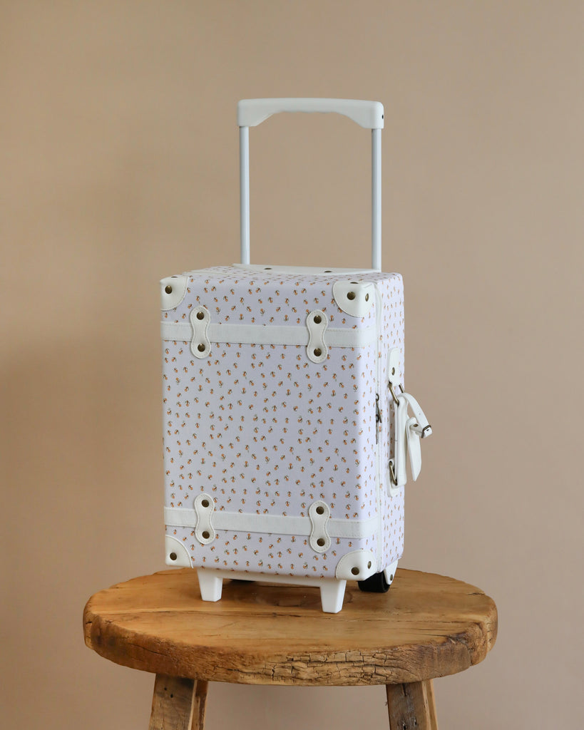 A stylish white Olli Ella See-Ya Suitcase - Leafed Mushroom with a subtle floral pattern and metal accents sits on a rustic wooden stool against a neutral beige background.