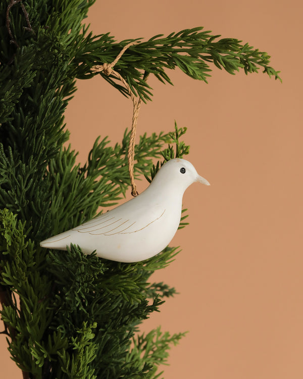 The Hand Carved Hanging Ornament - Dove Bird - 2.5"H is shown hanging from a green pine branch against a plain beige background.