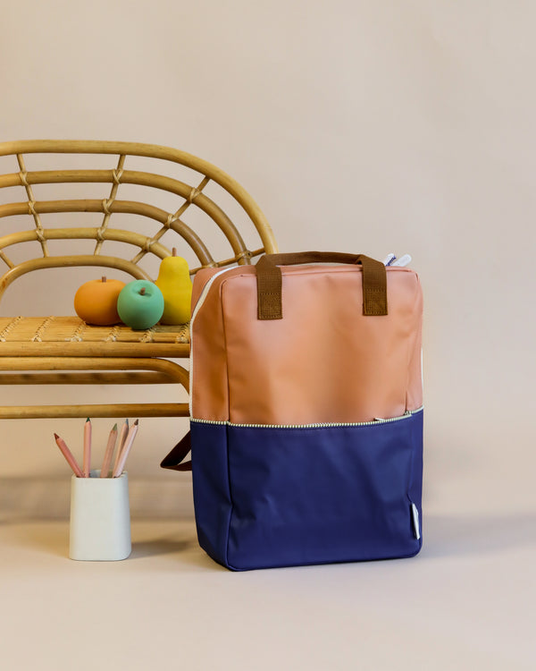 A Sticky Lemon Backpack Large with peach and navy-blue colors, featuring a YKK zipper, stands in front of a rattan chair holding colorful decorative objects on a neutral backdrop. A white cup with pencils is to