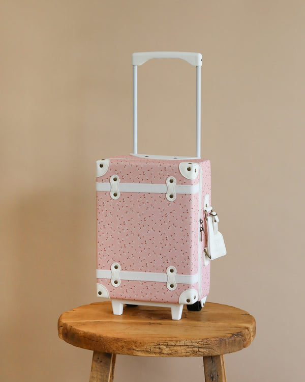 A pink Olli Ella See-Ya Suitcase - Pink Daisies with a white speckled pattern and metal corner details stands on an aged wooden stool against a neutral beige background. The suitcase handle is extended upwards.