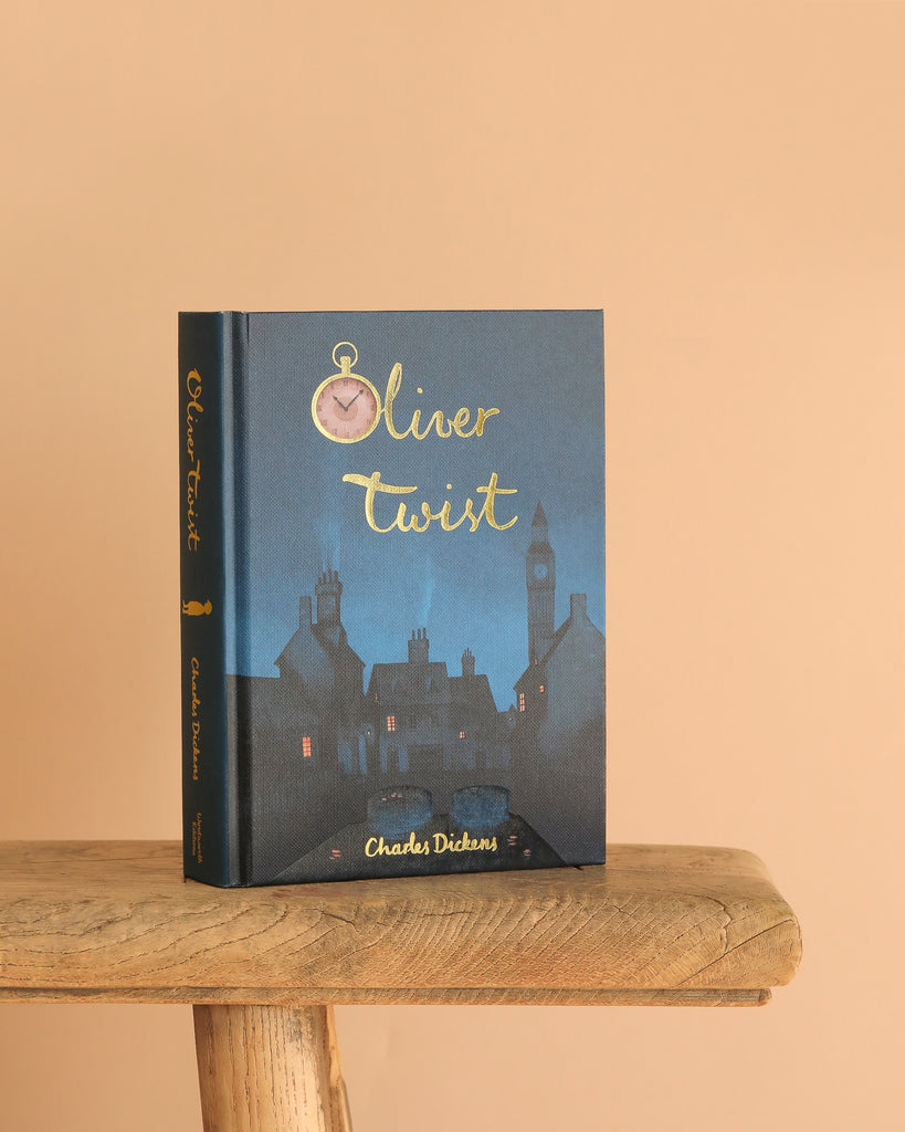 The Oliver Twist | Dickens | Collector's Edition | Hardcover Book stands on a wooden stool against a plain beige background, featuring an illustrated nighttime cityscape and a gold-scripted clock above the title.
