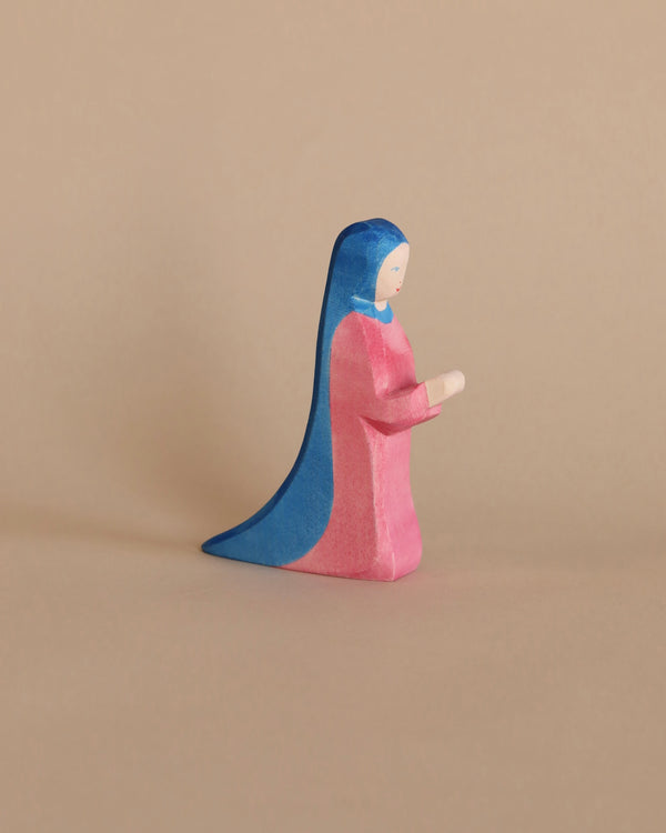 An Ostheimer Mary - Standing wooden figurine of a woman wearing a pink robe and blue headscarf against a plain beige background, depicted in a praying posture.