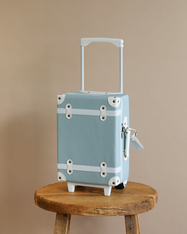 A light blue, vintage-style Olli Ella See-Ya Suitcase - Steel Blue with a silver handle extended, sitting on a wooden stool against a neutral beige wall. The suitcase has white trim and metal corners.