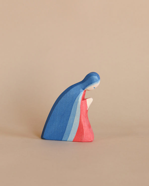 A handcrafted Ostheimer Mary figurine draped in a blue and red cloak against a plain beige background. The figure appears contemplative, with its head bowed.