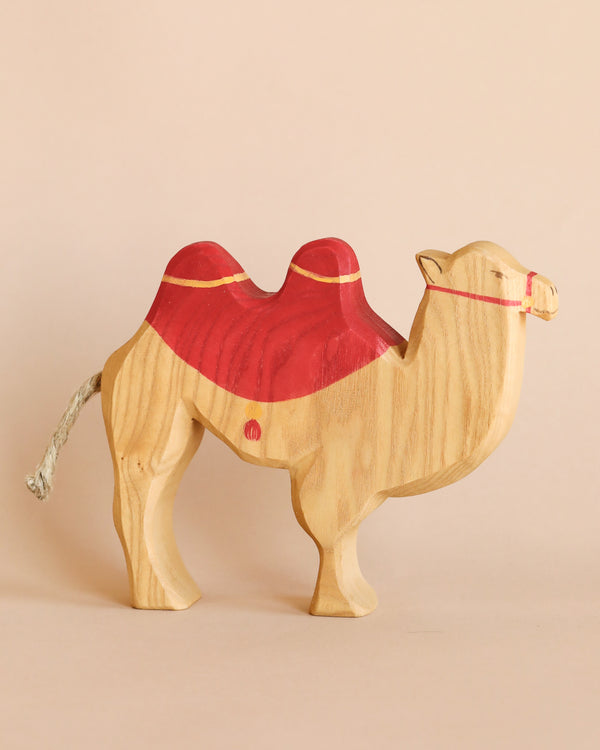 A Ostheimer Camel With Saddle figurine against a plain light pink background. Handcrafted as part of Ostheimer toys, the camel stands with a slight tilt, indicating movement, and has a detailed