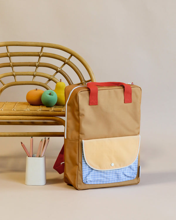 A beige Sticky Lemon Backpack Large with a blue and white checkered front pocket, crafted from waterproof nylon, stands next to a bamboo chair with colorful fruit and a cup of pencils on a neutral background.