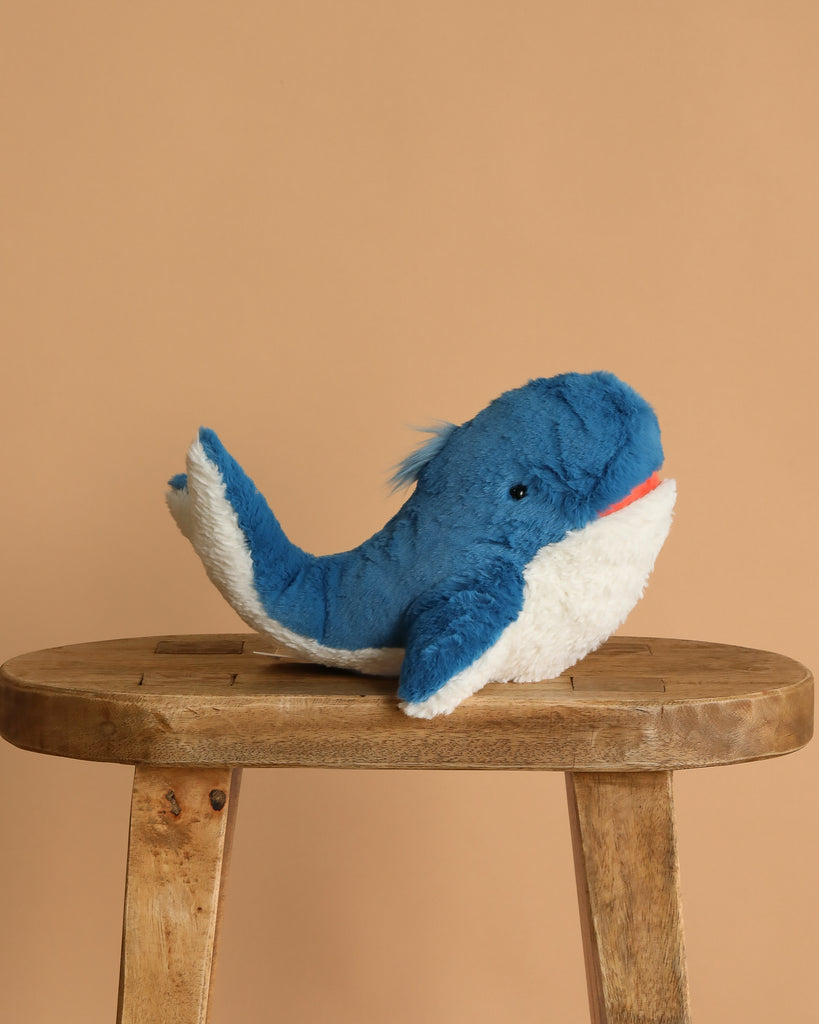 The Steiff Tory Blue Whale Plush Toy, 11 Inches, sits on a wooden stool against a plain beige background, making this adorable stuffed animal a charming addition to any nursery or playroom.