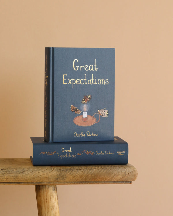 The Great Expectations | Collector's Edition | Hardcover stands upright on a rustic wooden stool, with another copy lying flat below it, set against a plain beige background.