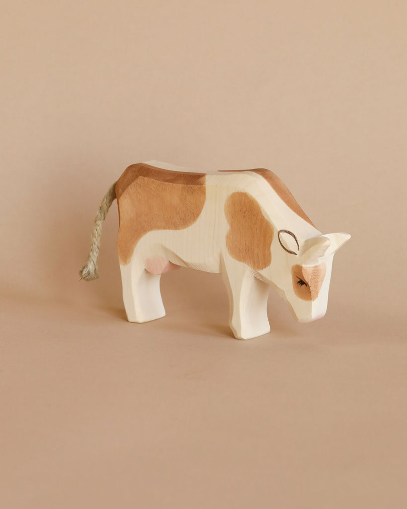 A Ostheimer Cow - Eating with patches of brown and white, standing against a soft beige background. The cow features detailed painting, including eyes and nostrils, and has a small rope tail.